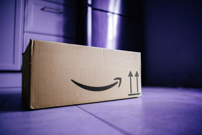 Una caja de cartón de Amazon con el icónico logotipo sonriente y una flecha que indica la orientación correcta para la entrega del paquete, sobre un fondo azul violeta.