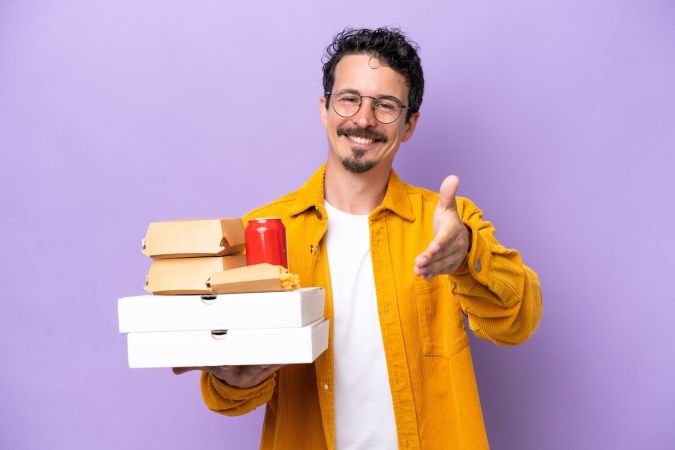 Joven caucásico sosteniendo comida rápida aislada sobre fondo morado estrechando la mano para cerrar un buen trato.