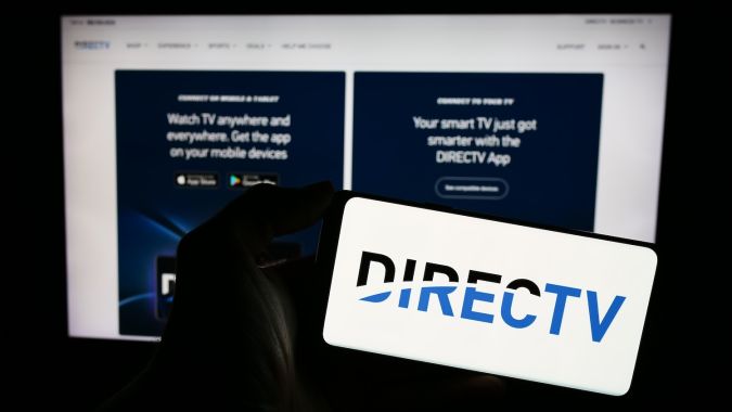 Persona que sostiene un teléfono móvil con el logotipo de la distribuidora de televisión estadounidense DirecTV LLC frente a la página web de la empresa.