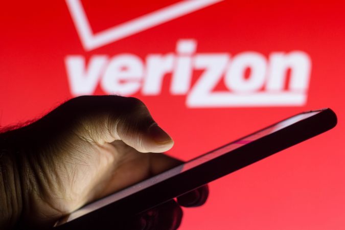 Se ve la silueta de una mano que sostiene un teléfono inteligente frente al logotipo de Verizon Wireless en el fondo.