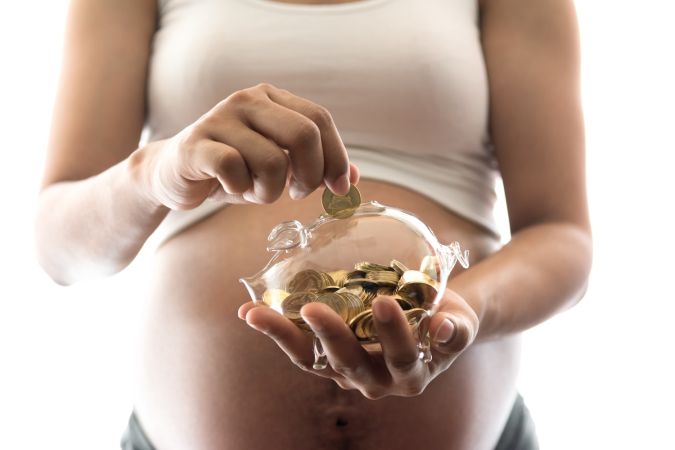 Mujer embarazada joven que deja caer una moneda de oro en el vaso de una alcancía para ahorrarle dinero a su bebé recién nacido aislado sobre fondo blanco.