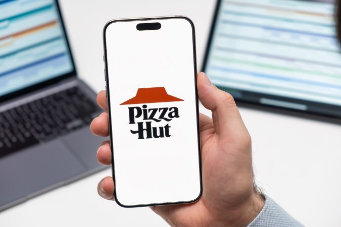 El logotipo de Pizza Hut en la pantalla del teléfono inteligente en la mano de un hombre y una computadora portátil en el fondo.